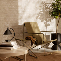 Modern olive green chair with minimalist black lamp and round beige table, set against a textured light wall.