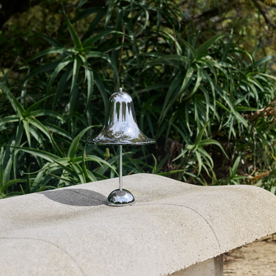 Shiny silver bell with a smooth, reflective surface, set against lush green foliage. Perfect for decorative use.
