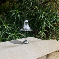 Shiny silver bell with a smooth, reflective surface, set against lush green foliage. Perfect for decorative use.