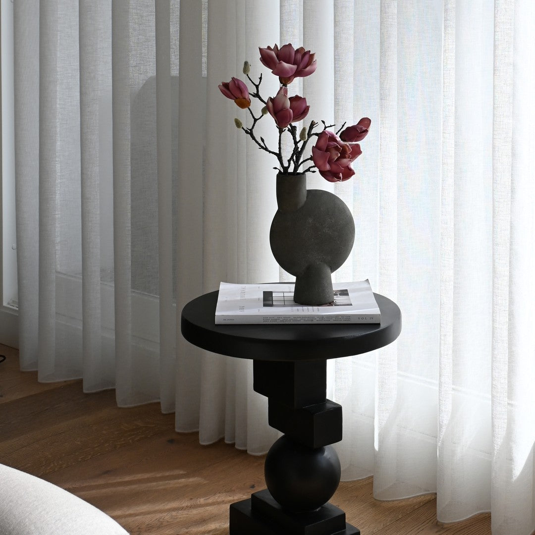 "Elegant black vase with purple flowers on a black table, surrounded by light gray curtains and wooden flooring."