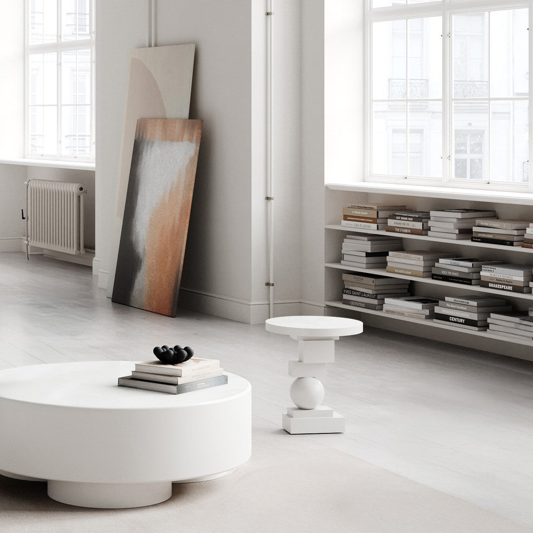 Modern living room with a white round table, warm-toned abstract art, and neutral colored books on shelves.