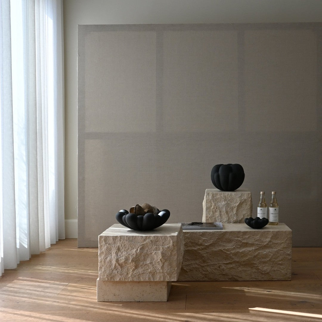 Black ceramic vases on textured stone pedestals against a neutral beige background, highlighted by soft natural light.