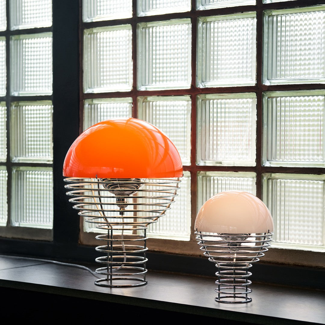 "Orange and white dome lamps with sleek metal wire bases, illuminated against a backdrop of textured glass windows."