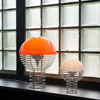 "Orange and white dome lamps with sleek metal wire bases, illuminated against a backdrop of textured glass windows."
