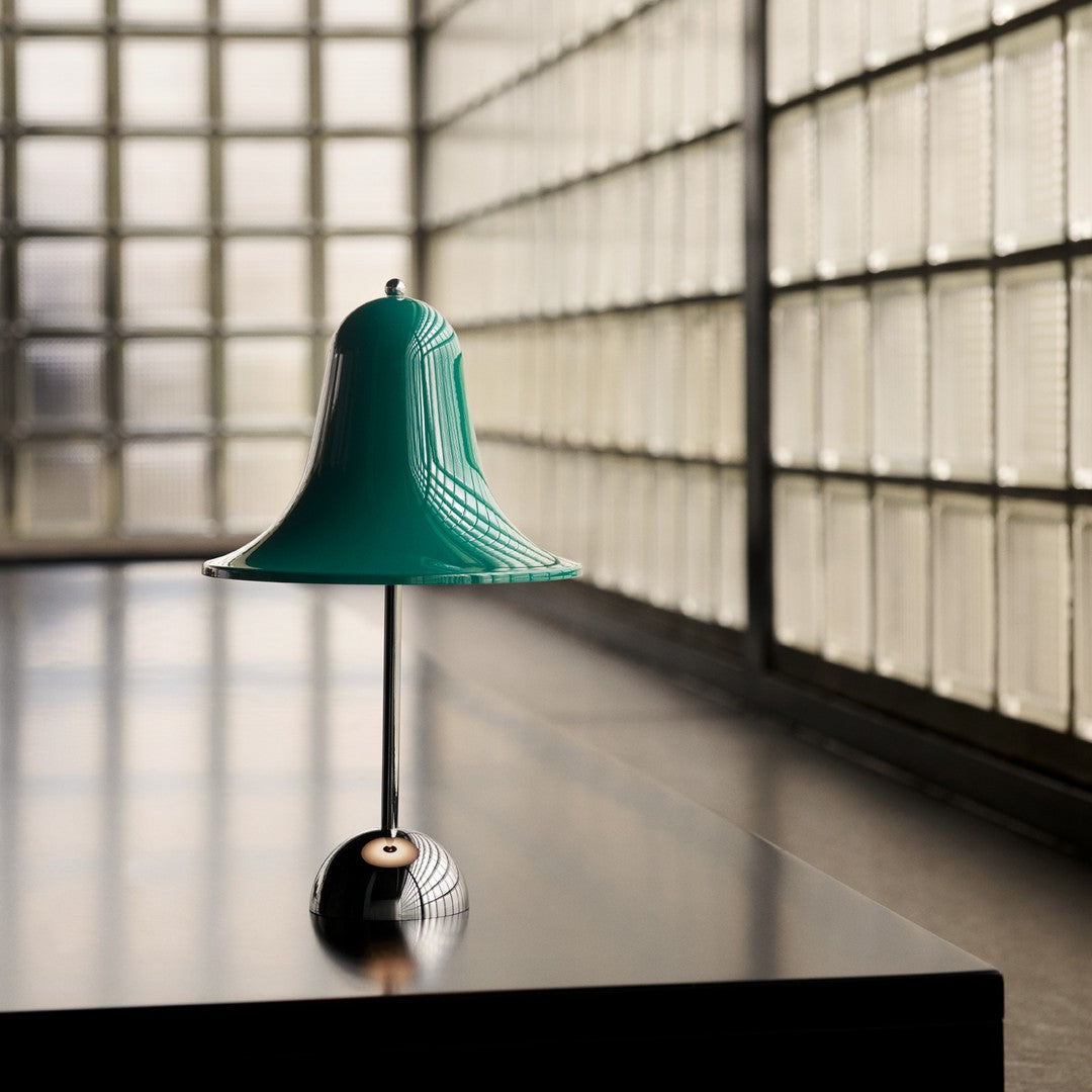 Teal bell-shaped lamp with a glossy finish, standing on a black base against a textured glass backdrop.