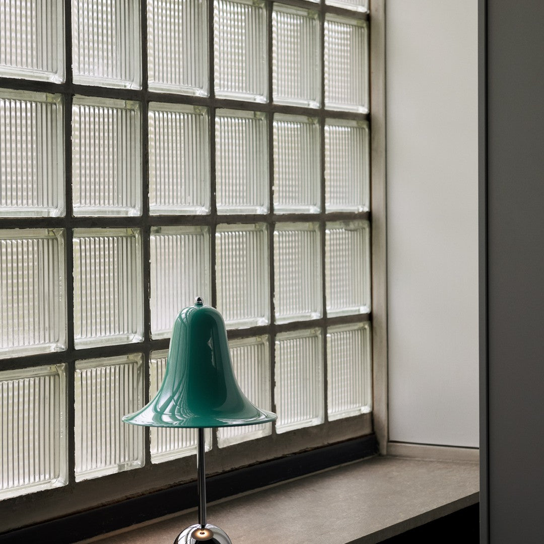 Teal bell-shaped lamp with glossy finish, contrasting against textured glass block window. Elegant modern decor.