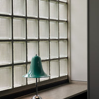 Teal bell-shaped lamp with glossy finish, contrasting against textured glass block window. Elegant modern decor.