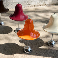 Colorful bell-shaped stools in orange, burgundy, cream, and brown with shiny chrome bases on a sandy surface.