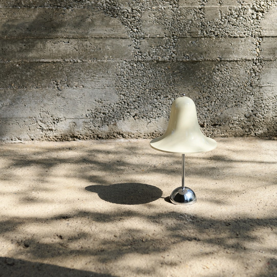 Cream-colored bell-shaped lamp on a metallic base, casting soft shadows on a textured sandy surface.