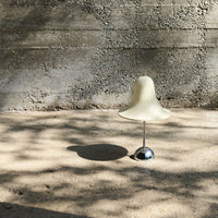 Cream-colored bell-shaped lamp on a metallic base, casting soft shadows on a textured sandy surface.