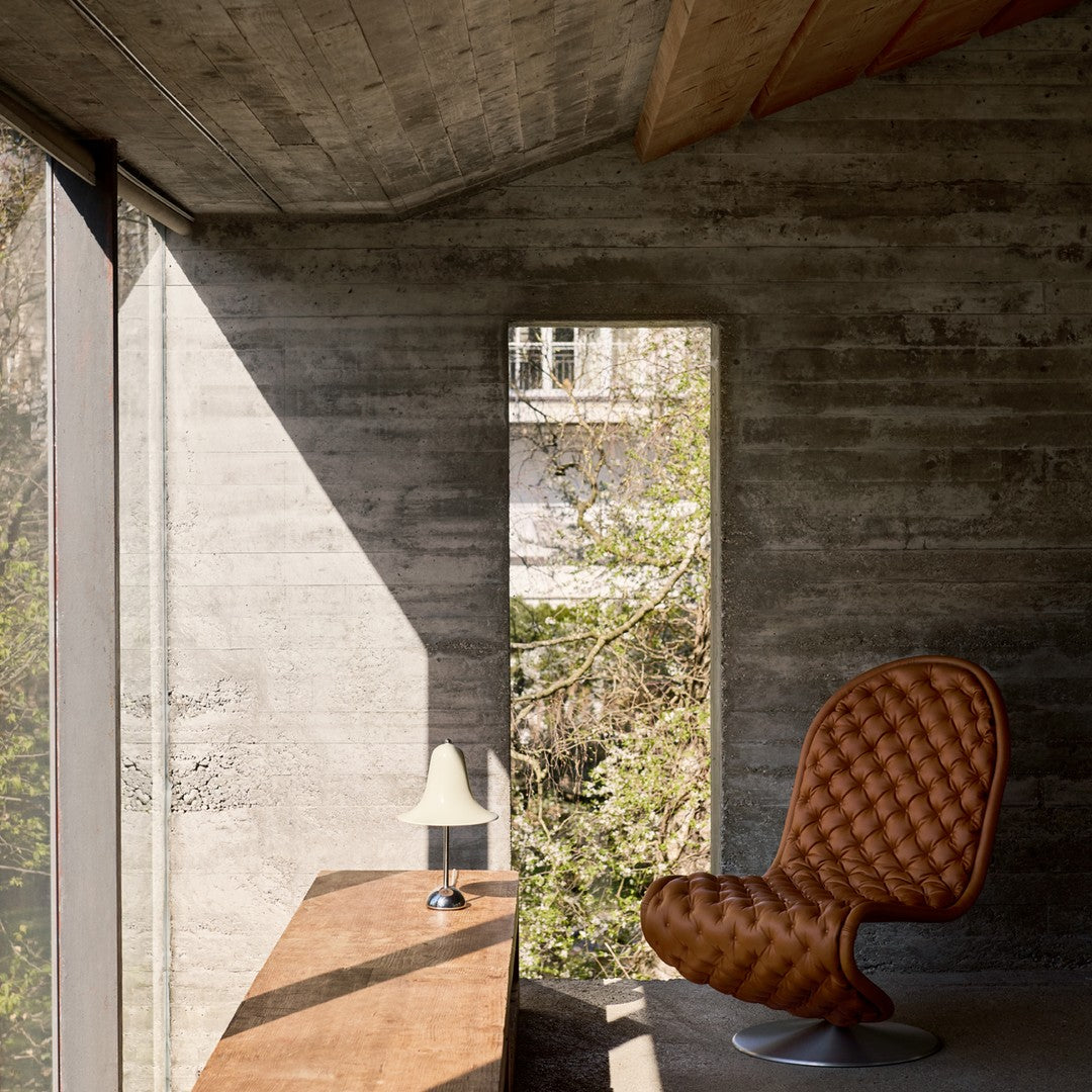 Brown tufted chair with smooth silver base, set against a sleek concrete wall and bright natural light.