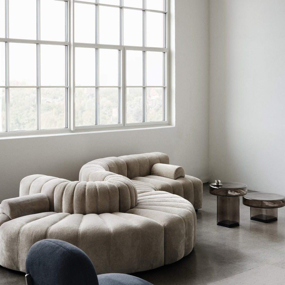 Beige curved sofa with plush texture, complemented by dark glass accent tables, in a bright minimalist room.
