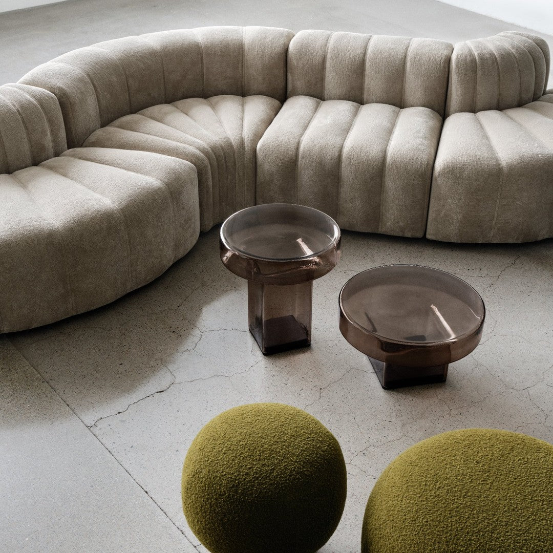 Beige curved sofa with textured fabric, brown glass tables, and green textured round ottomans on a gray floor.