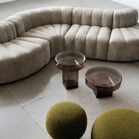 Beige curved sofa with textured fabric, brown glass tables, and green textured round ottomans on a gray floor.