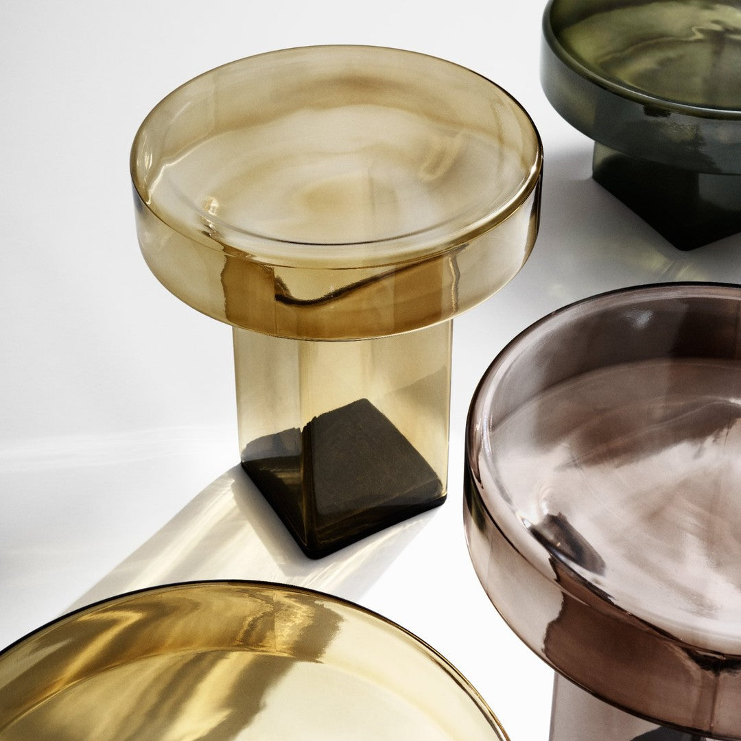 Transparent glass tables in amber, olive green, and smoky brown hues with smooth, glossy finishes.