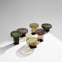 Set of five modern glass tables in smoky green, amber, and brown hues with geometric bases.