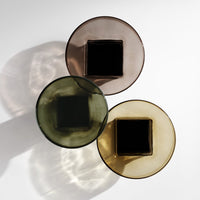 Three decorative glass bowls in amber, olive green, and smoky brown with square bases and smooth finishes.