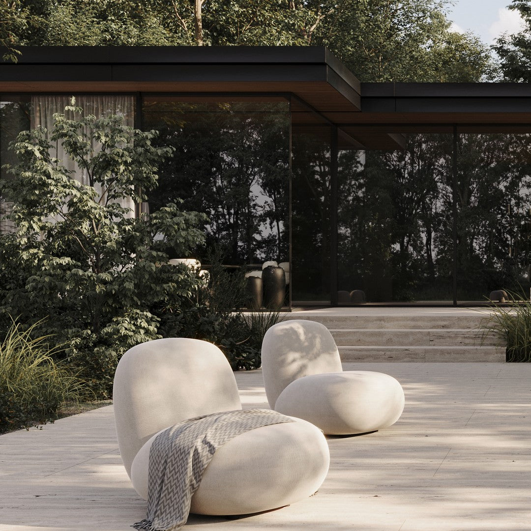 Beige lounge chairs with soft contours, paired with a textured gray throw, set against lush greenery and modern architecture.