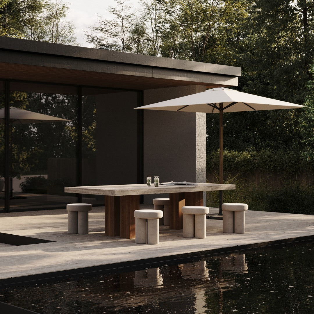 Modern outdoor dining set with a gray concrete table, light beige stools, and a white umbrella against lush greenery.