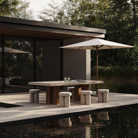Modern outdoor dining set with a gray concrete table, light beige stools, and a white umbrella against lush greenery.