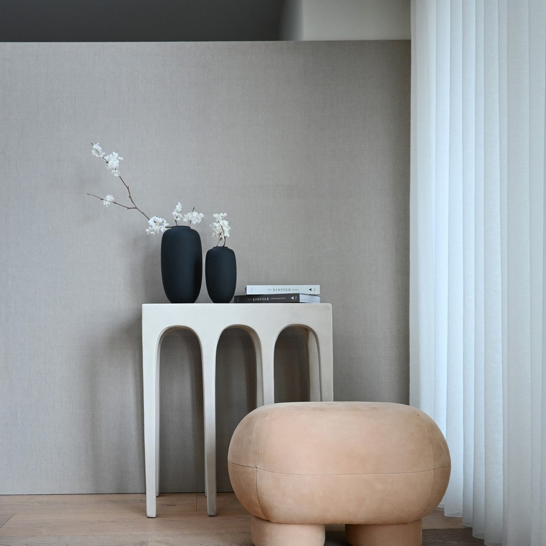 Minimalist beige backdrop with black vases and a soft tan pouf, accented by white floral decor and layered textures.