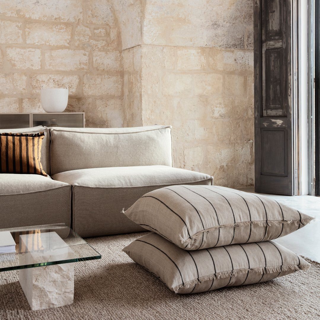 Beige sofa with striped cushions in earthy tones, set against rustic stone walls and a light-colored woven rug.