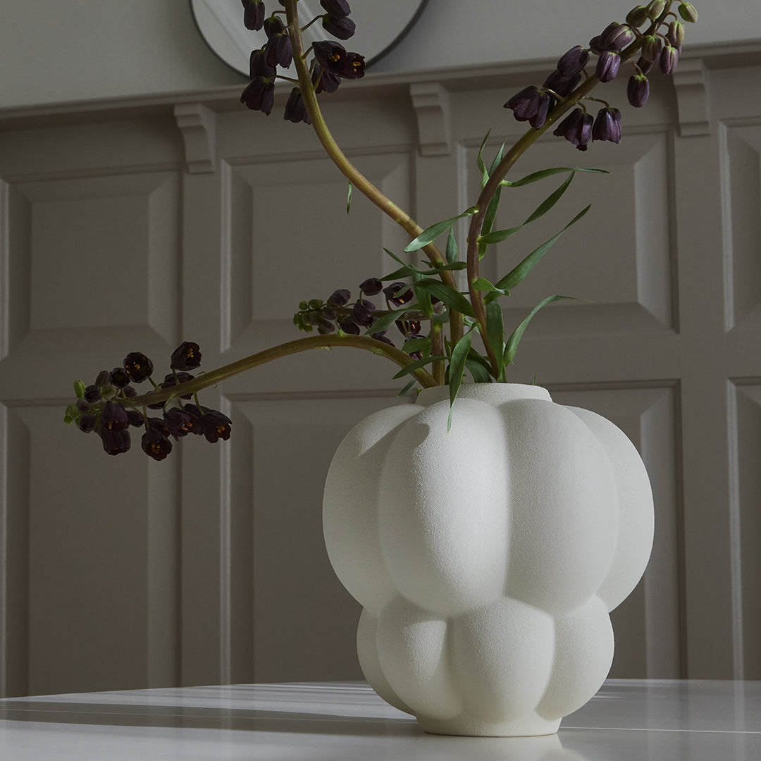 White round vase with subtle ribbed pattern, showcasing dark purple flowers against a light background.