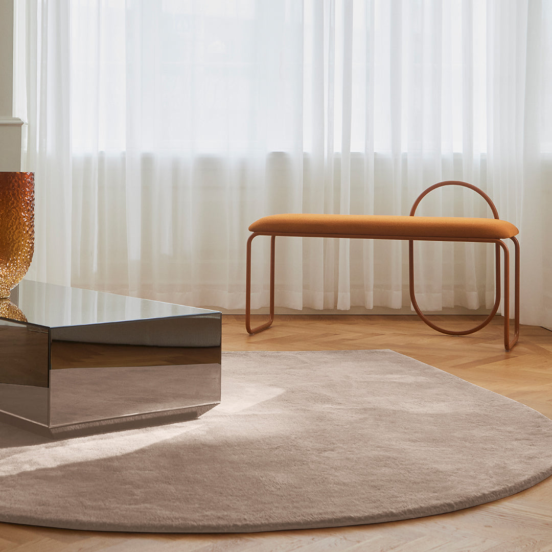 "Soft beige circular rug with a sleek metallic coffee table and vibrant orange bench against a bright, airy setting."