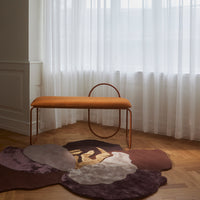 "Orange upholstered bench atop a floral-patterned rug in soft purples and whites, set against sheer white curtains."