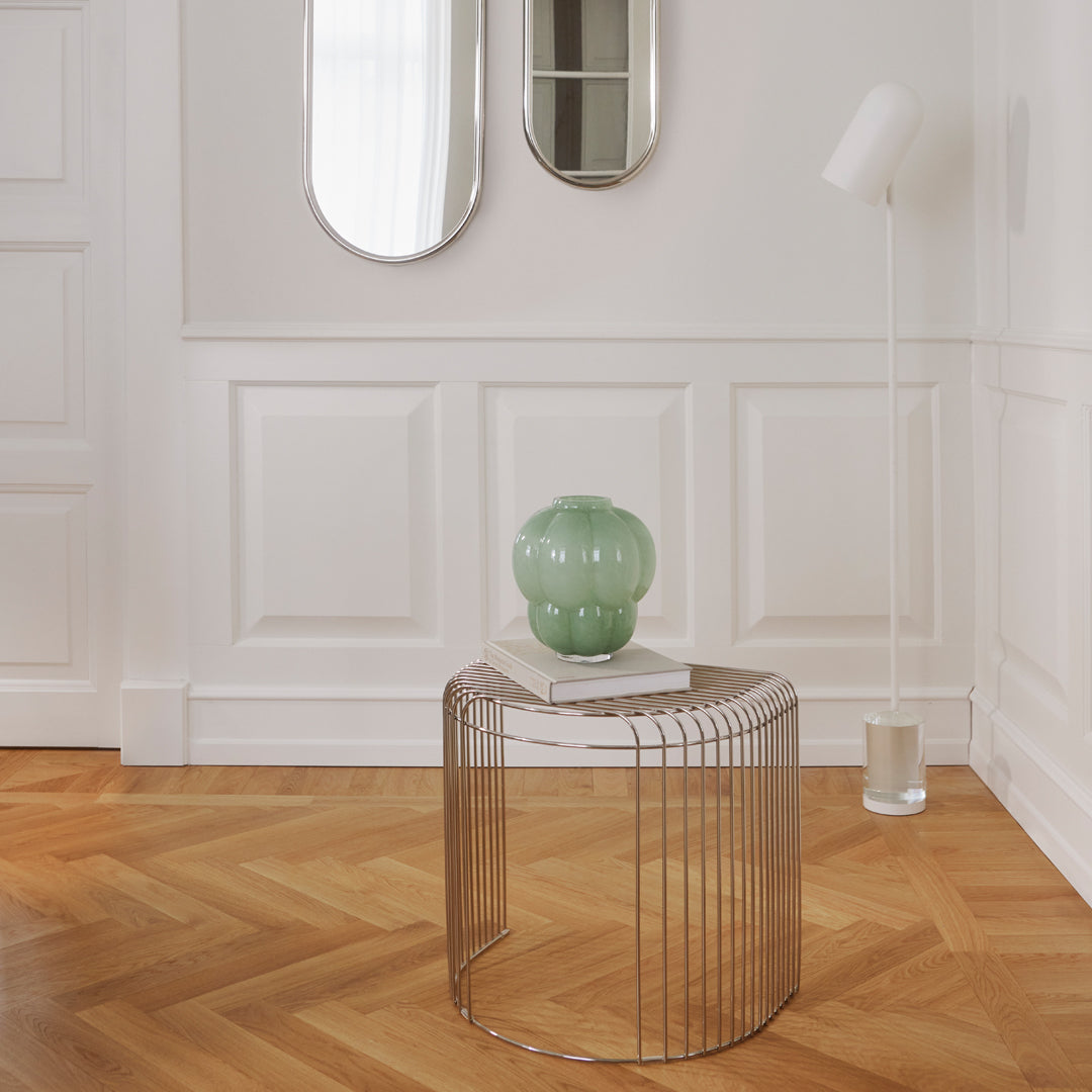 Modern striped wire table with a round mint green vase on a light wood herringbone floor and elegant white walls.
