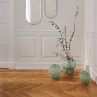 "Three green glass vases with a bubble pattern against a white wall and herringbone wood flooring."
