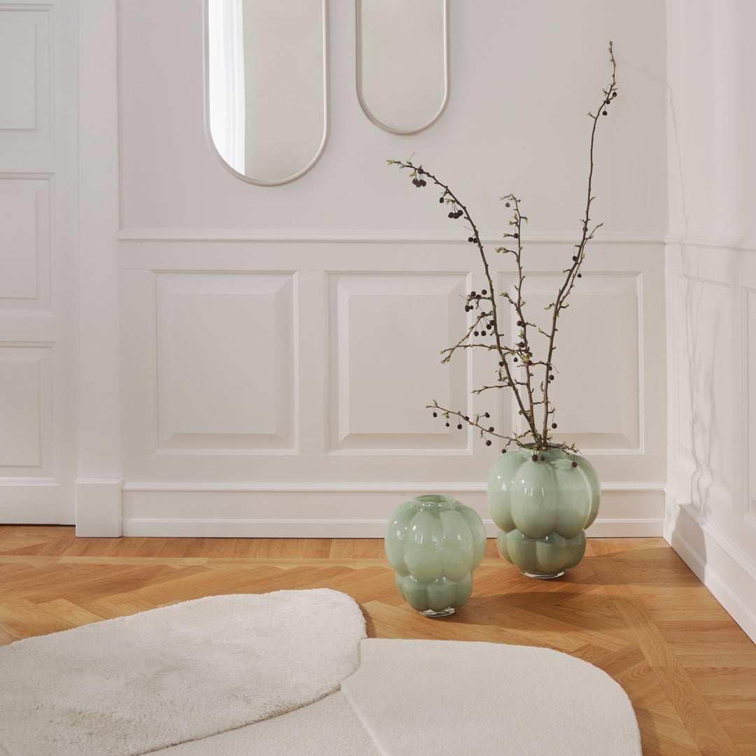 Green glass vases with smooth, rounded shapes, set against a light wood floor and white textured walls.