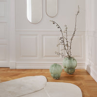 Green glass vases with smooth, rounded shapes, set against a light wood floor and white textured walls.