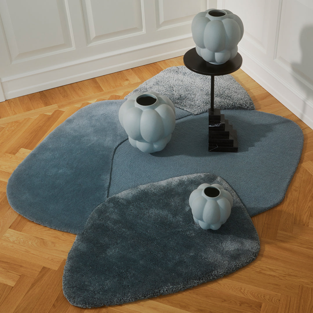 "Soft blue rugs with varying shades and organic shapes, complemented by light blue vases and a black pedestal table."