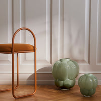 "Light green glass vases with a textured, rounded design next to a warm orange chair against a neutral backdrop."