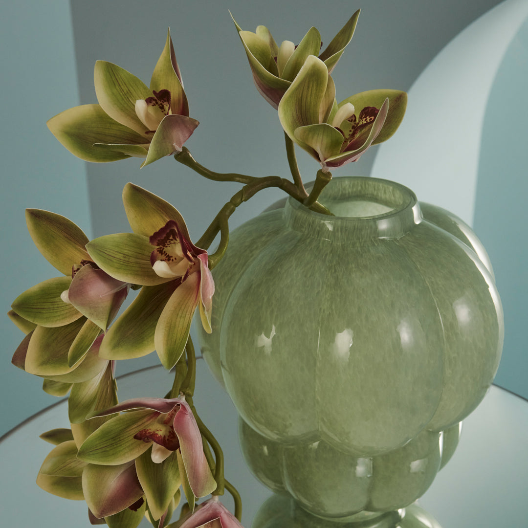 Green glass vase with a glossy finish, adorned with pink and green orchid flowers. Elegant and modern design.
