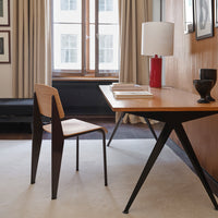 Wooden desk with light brown finish, black metal legs, a red lamp, and minimalist decor. Soft neutral tones in the background.