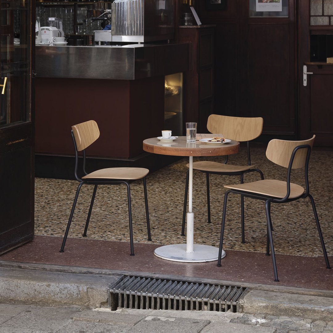 Outdoor café set featuring a round wooden table and two simple black and natural wood chairs against a rustic backdrop.