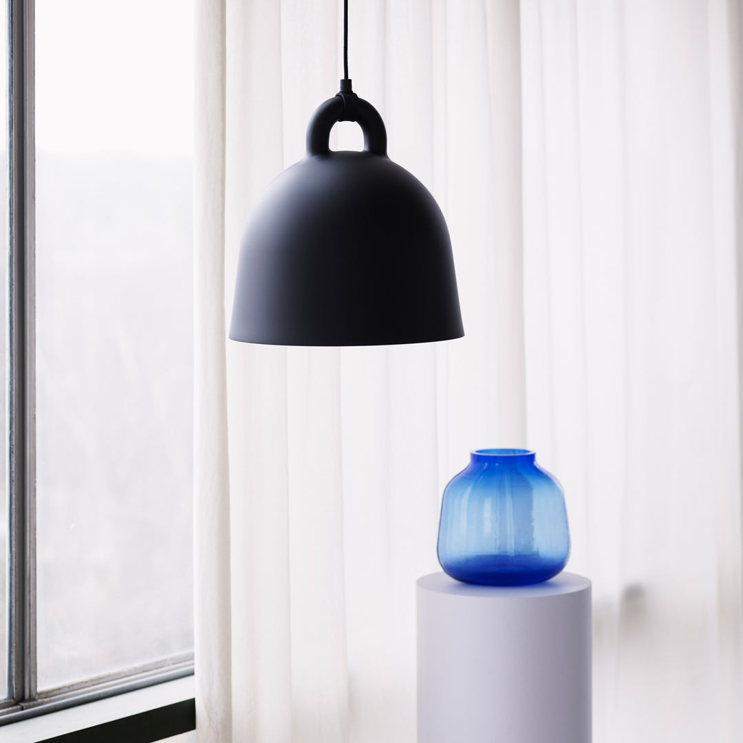 Modern black pendant lamp and a round blue decorative vase on a white pedestal, set against sheer curtains.