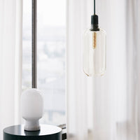 Clear glass pendant light with a sleek black cord, complemented by a smooth white lamp base and neutral curtains.