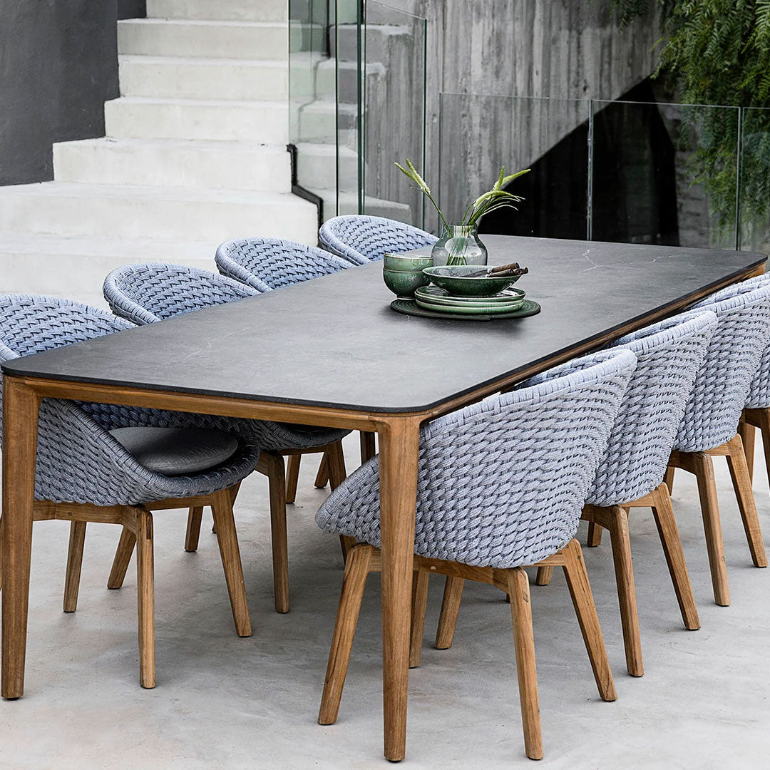 Gray woven chairs with wooden legs around a sleek black table, accented by green plates and a glass vase.