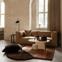 Modern beige sofa with earthy-toned cushions, a round white coffee table, and a geometric-patterned brown rug.