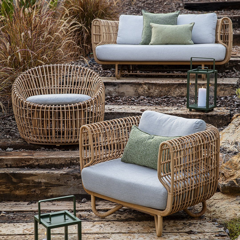 Natural rattan furniture set featuring grey cushions and a green accent pillow, set against a rustic outdoor backdrop.