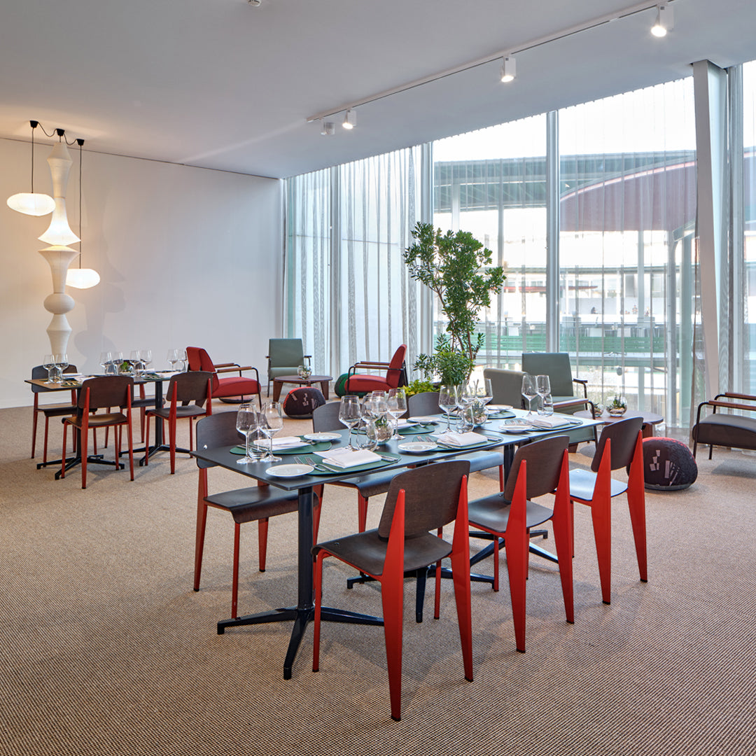 Modern dining room with red and black chairs, glass table, and neutral-toned carpet. Minimalistic design and natural light.