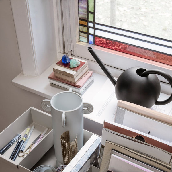 Modern black kettle with glossy finish beside colorful stacked books and a white geometric container on a bright windowsill.