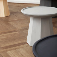 Gray geometric side table on wooden floor, with black and light wooden stools in the background. Modern minimalist design.