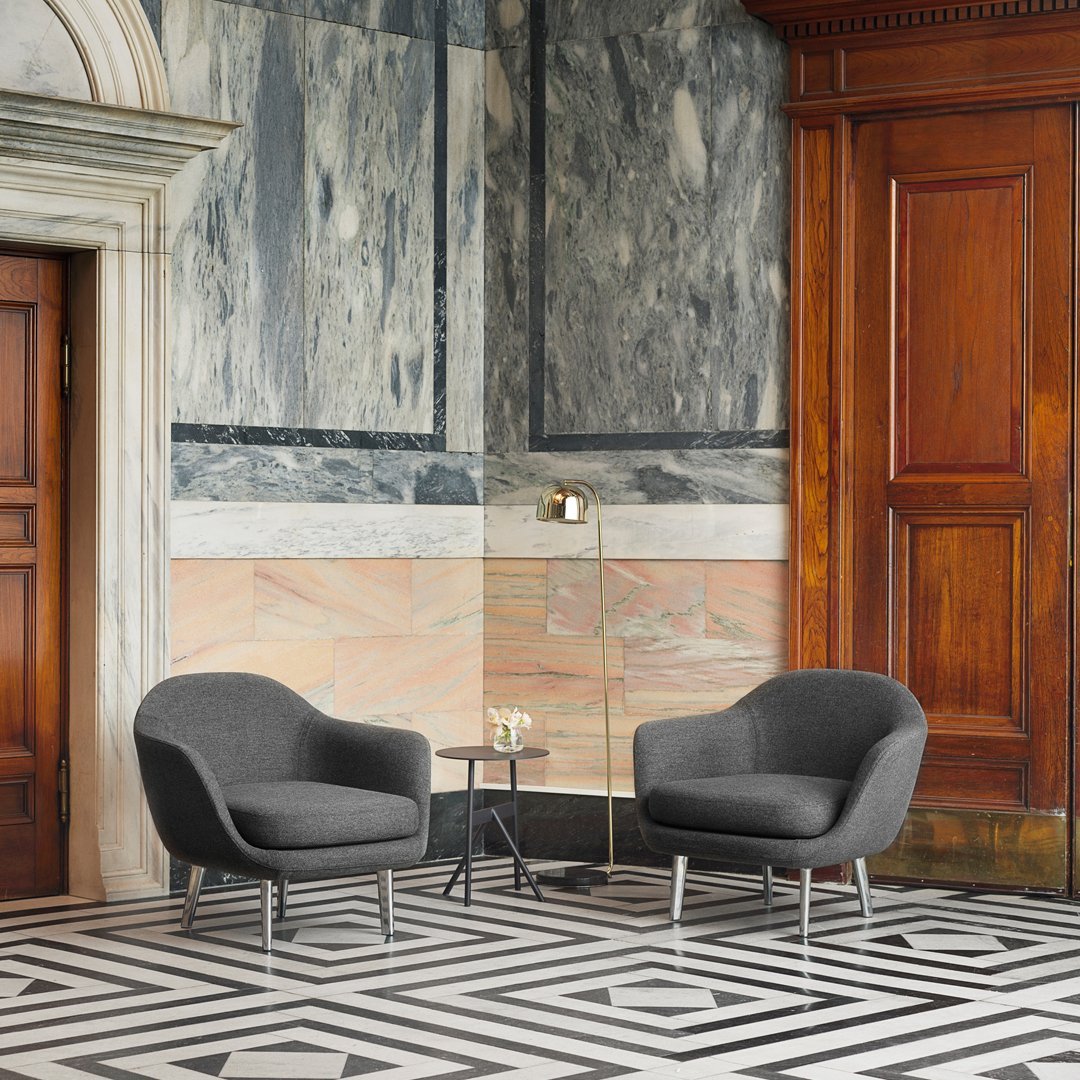 Two gray armchairs on a bold black-and-white patterned floor against a marble and wooden backdrop.