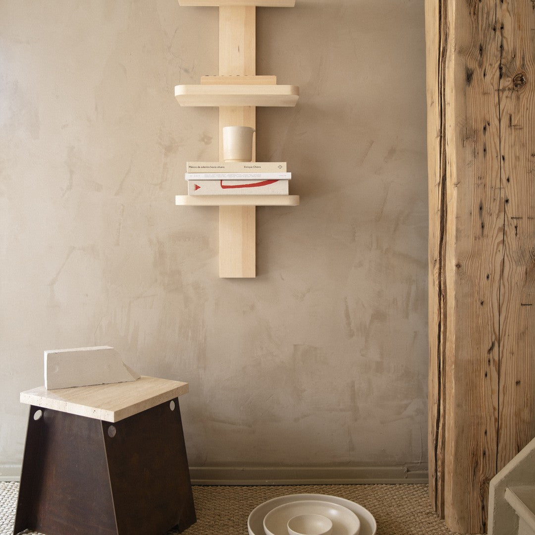 Light wood shelving unit against a neutral textured wall, with a dark wood stool and white circular plates below.