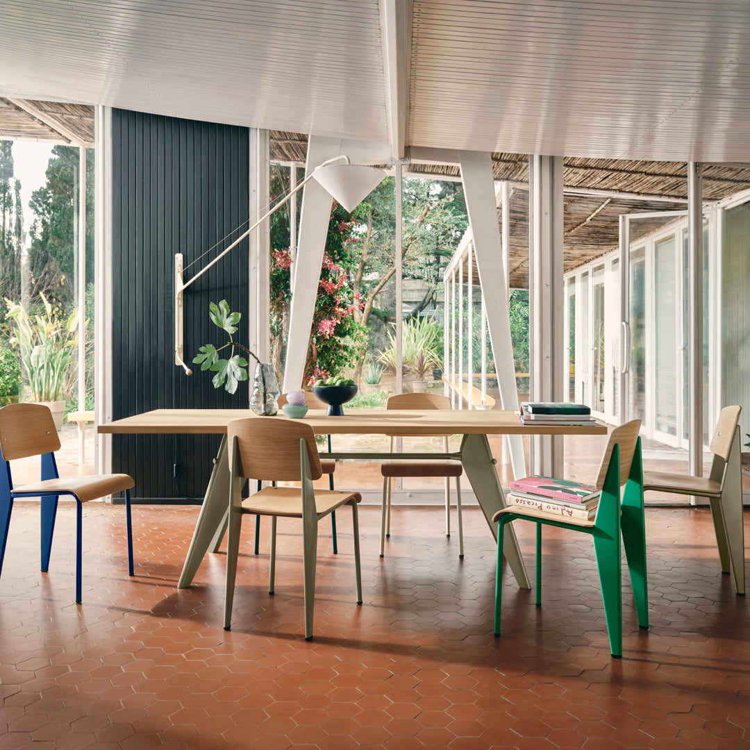 Modern dining room with a light wood table, colorful chairs in blue, green, and natural wood tones, and a warm terracotta floor.