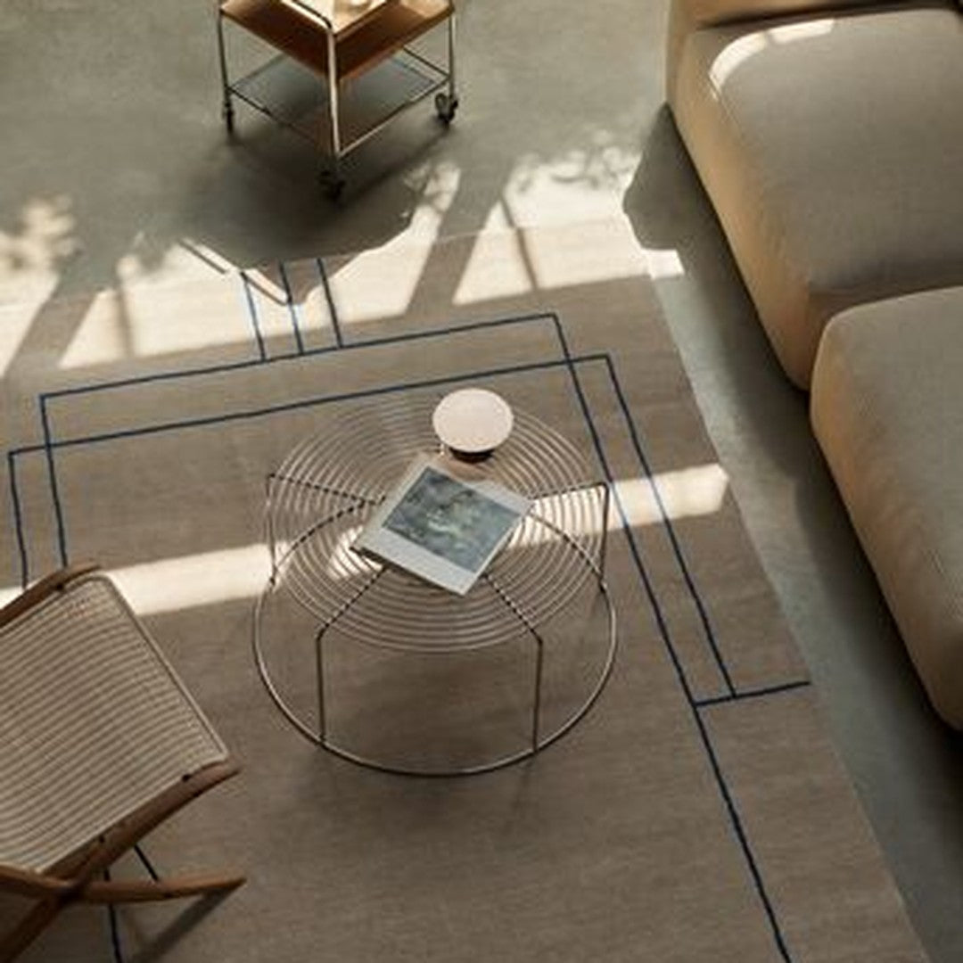Neutral beige area rug with blue geometric lines, complemented by a minimalist wire coffee table and soft, light furnishings.